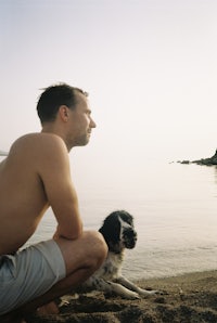 a man crouching on the sand next to a dog